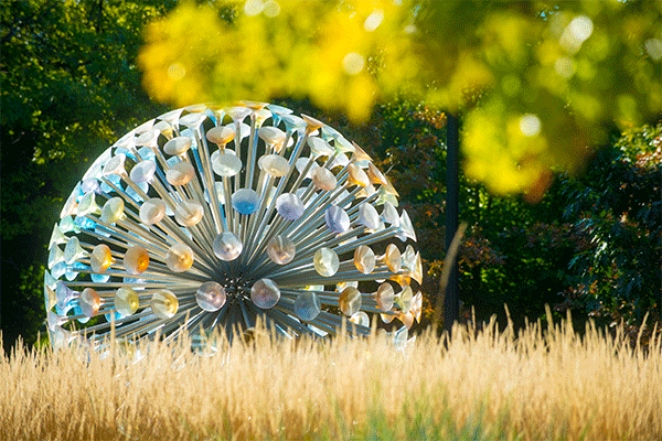 Photo of a large, circular structure made up of mall colorful disks suspended by metal posts installed in a round core, with brown native grass in the foreground and green foliage from a tree in the top right corner. 