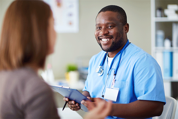 Black male with short hair, beard and mustache wearing blue nursing scrubs and a nametag and holding a clipboard smiles and talks to a female patient with her back to the camera in a medical office setting.   