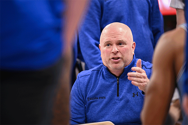 Middle-aged white male wearing a blue zipped-up shirt and talking to basketball players with their back to the camera.