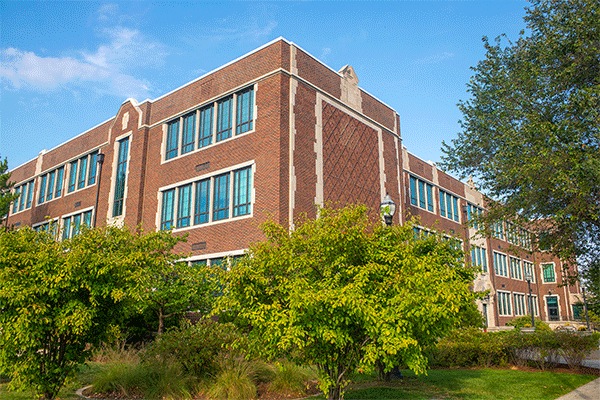 Exterior image of a large, multi-story brick building that houses the Bayh College of Education with green trees obscuring the lower half of the image with a blue sky.  