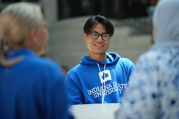 A male student of Asian descent with short dark hair, glasses, and a blue hoodie with Indiana State University in white lettering smiles while sitting at a table. He talks to two female students with their backs to the camera.  