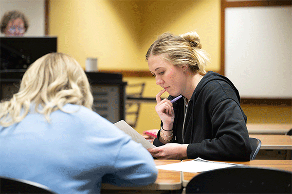 Blonde female student looking over a paper in class with another student whose back is to the camera and a professor visible in the background.