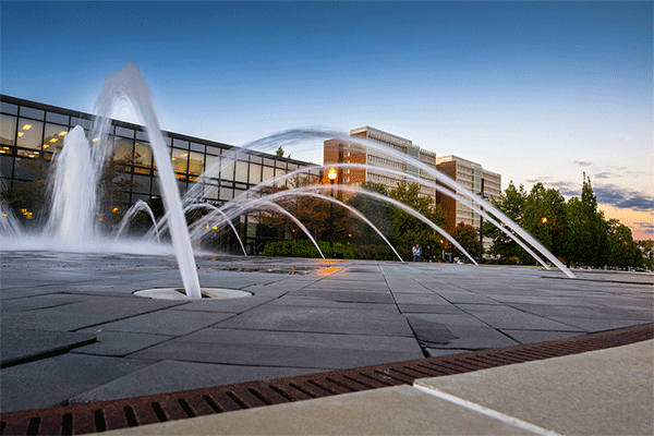 Low angle of a fountain with streams of water in the foreground and the Welcome Center and Sycamore Towers and blue sky in the background. 