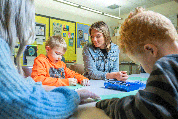 A blonde teaching student with short hair and wearing a blue jean jacket is seated at a table in a classroom. She watches as a young blond boy in an orange sweatshirt seated next to her creates a drawing on green construction paper. In the foreground, another young boy with short red hair works on a drawing, while another female teaching student with long blonde hair and wearing a blue sweater holds the paper in place for him. 