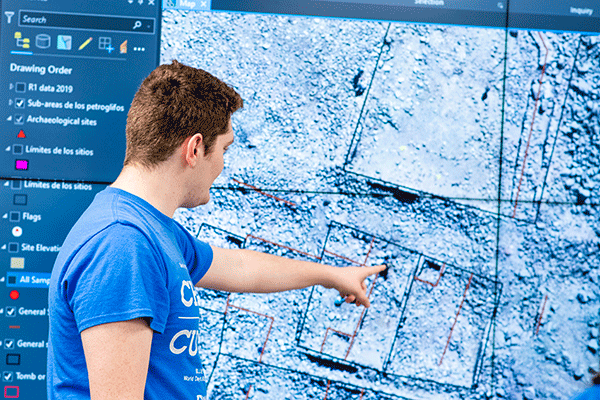 A white male student with short brown hair stands in front of a large digital screen. He wears a blue T-shirt with “Change the Culture” in large white lettering on the front. He also wears denim jeans. He is pointing to the screen with his left hand. The screen displays a close-up view of grey dirt. Black squares are drawn on the screen. 