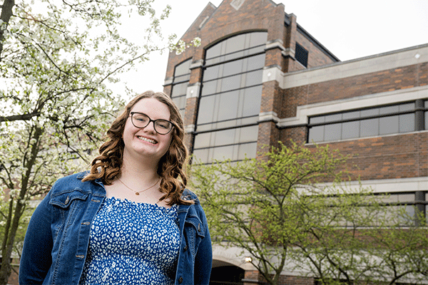 Outdoors photo of a white female student with glasses and long brown hair standing and smiling at the camera with the brick entrance of the Bailey College of Engineering and Technology in the background. Green trees are visible in the background. 