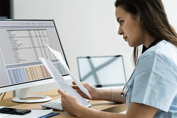 Female wearing scrubs and seated at a desk reviewing papers and a spreadsheet on a desktop in the background. 