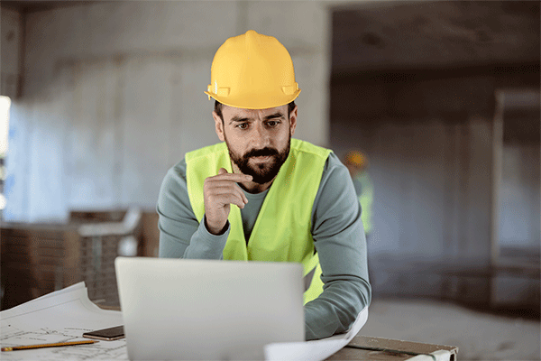 Middle-aged male with dark beard and mustache and wearing a hard had and yellow safety vest while sitting at a table in front of a laptop in a construction zone. 