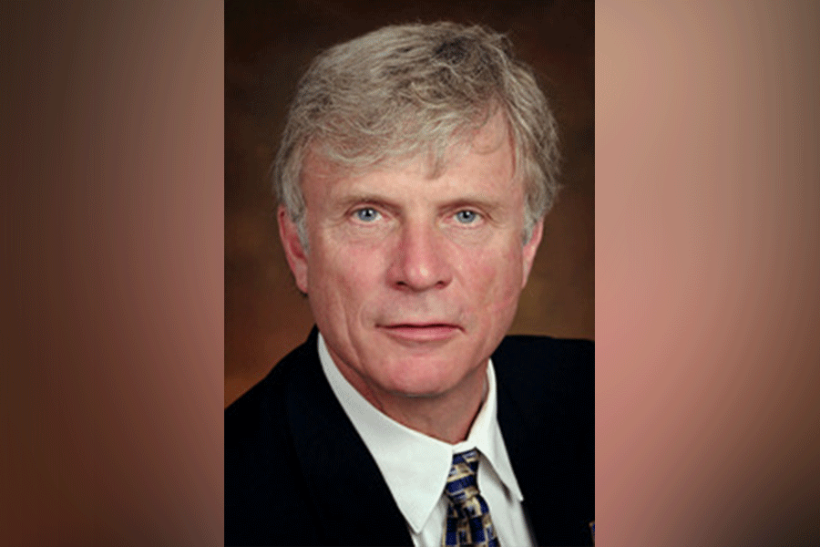 A man (President Lloyd W. Benjamin III) with short greying hair. He looks straight ahead. He wears a black suit with a white shirt and a tie. A light brown border is around the photograph. 