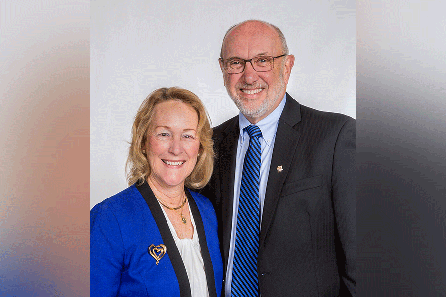 A photograph of President Daniel J. Bradley with his wife, Cheri Bradley. On the left, Cheri is a white female with shoulder-length blonde hair. She wears a blue blazer with a white shirt. On the right is President Daniel J. Bradley, a white male with short grey hair and a matching grey beard. He wears glasses, a black dress suit, a light blue shirt, and a blue-and-black striped tie. A grey border is around the photograph. 