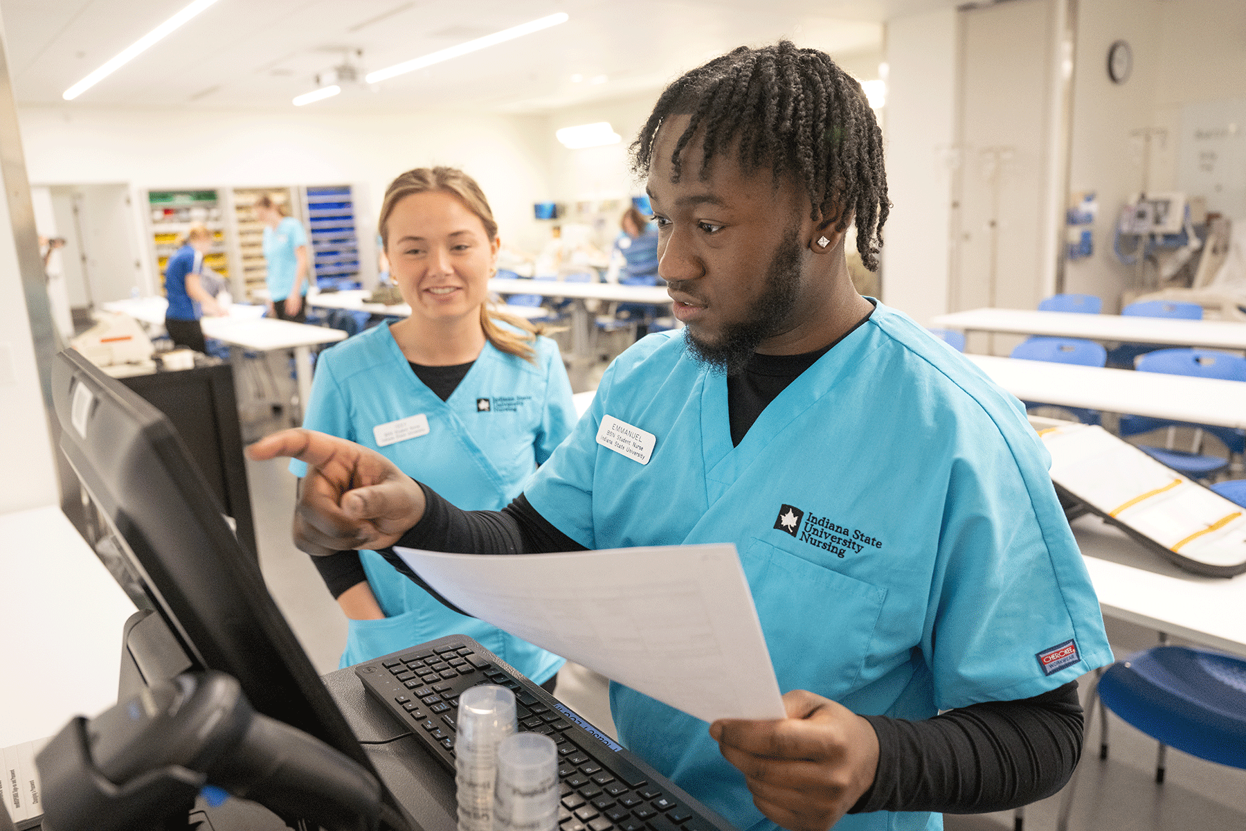 A male and a female nursing student, smiling and wearing ISU scrubs, gain knowledge and skills in one of the Nursing Learning Resource Center skill labs at Indiana State University. 