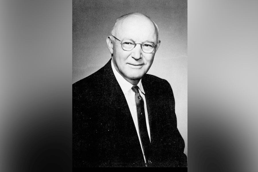 A black-and-white photograph of a man (President Raleigh Warren Holmstedt). He is bald and wears glasses. He looks ahead and to the left. He wears a suit with a white shirt and tie. A grey border is around the photograph. 