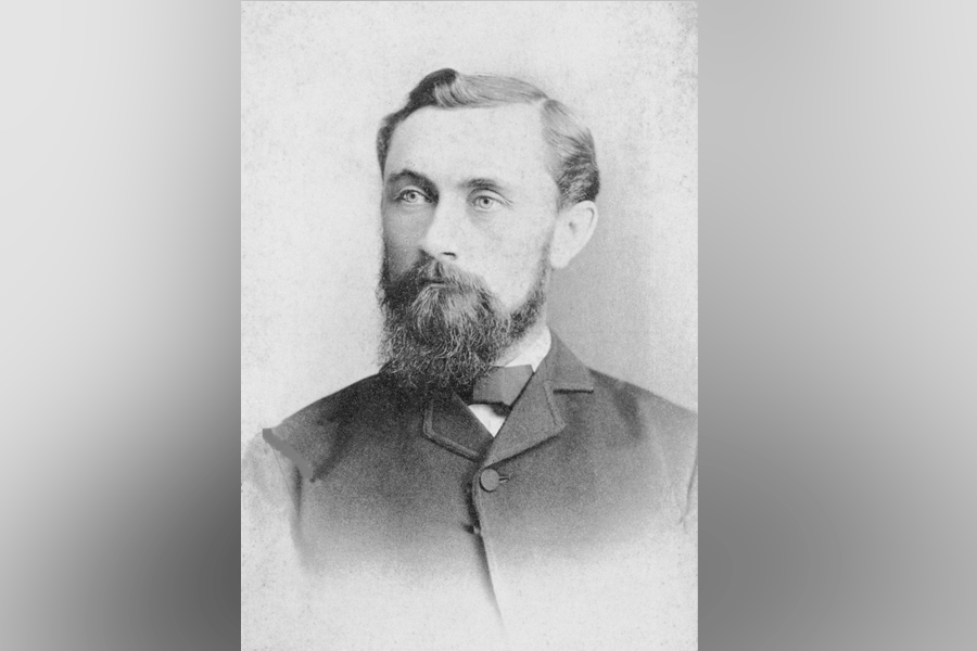 A black-and-white photograph of a man (President William Albert Jones) with short hair and a full beard and mustache. He looks ahead and to the left. He wears a button-up suit with a bowtie. A grey border is around the photograph. 