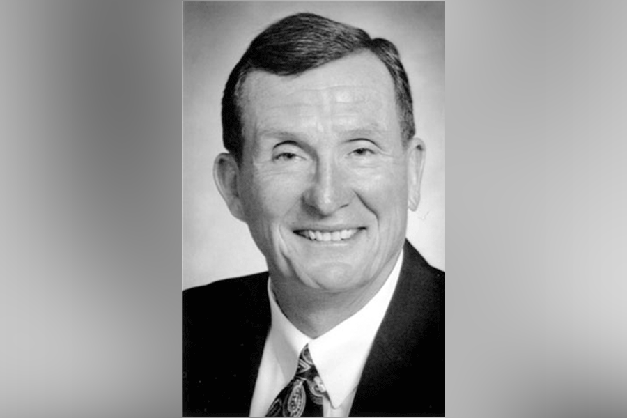 A black-and-white photograph of a man (President John William Moore) with short hair. He smiles as he looks straight ahead. He wears a suit with a white shirt and tie. A grey border is around the photograph. 