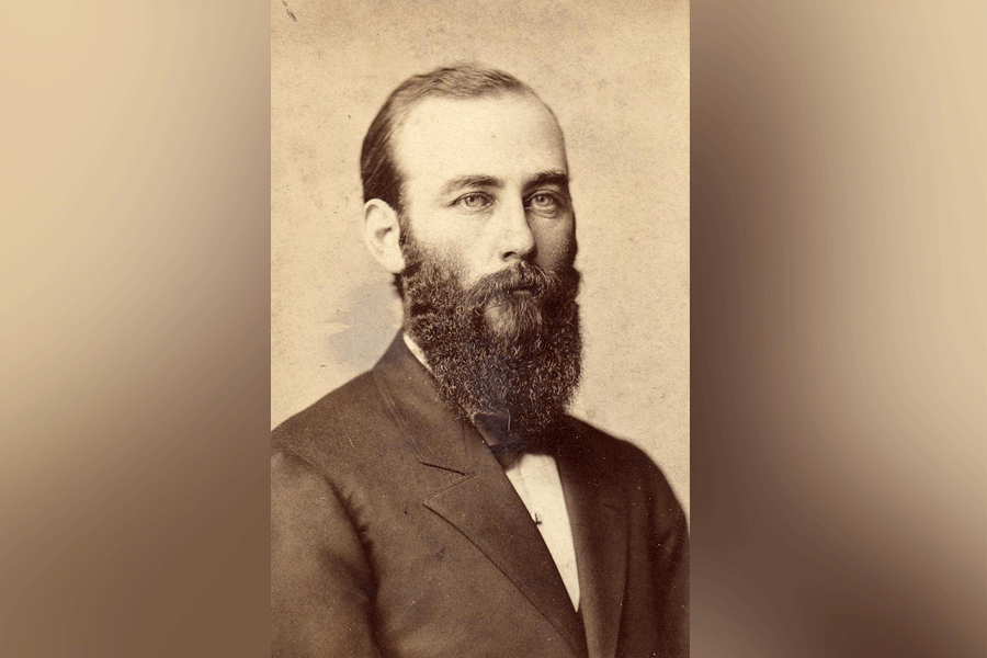 A black-and-white photograph of a man (President William Wood Parsons) with short hair and a full, long beard and mustache. He looks ahead and to the right. He wears a suit with a white undershirt and a bowtie. The photograph appears to have a light brown tint. A light brown border is around the photograph.  