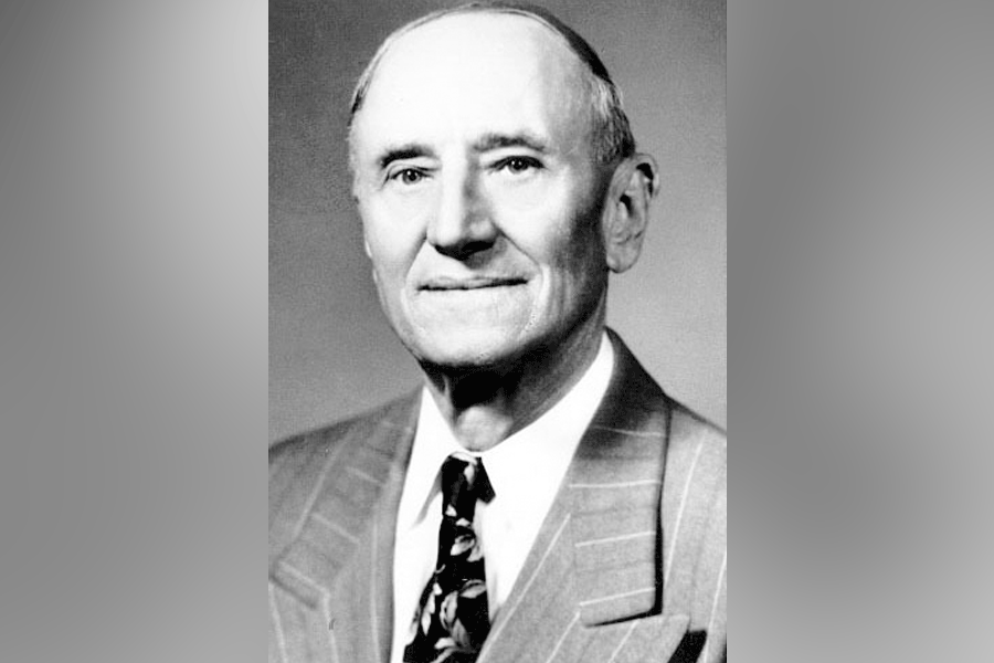 A black-and-white photograph of a man (President Ralph Noble Tirey) with short hair. He looks straight ahead. He wears a suit, a white shirt, and a tie. A grey border is around the photograph. 