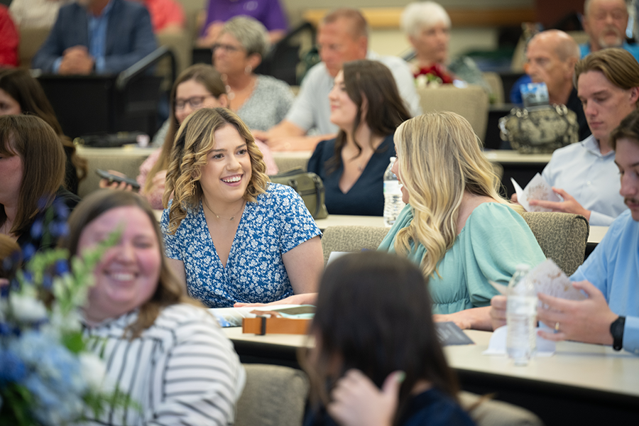 A smiling, blonde female student, wearing a white dress with black line patterns on it, stands to the left of a smiling female faculty member with dark hair who is wearing a white and lavender dress with a grey and lavender scarf. They both look forward while posing for a photo in a classroom setting with the photographer visible on the right side of the frame and out of focus.  