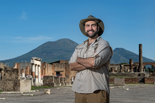 A male professor stands with arms crossed facing the camera in an open area within the ruins of Pompeii with Mt. Vesuvius visible in the background. 