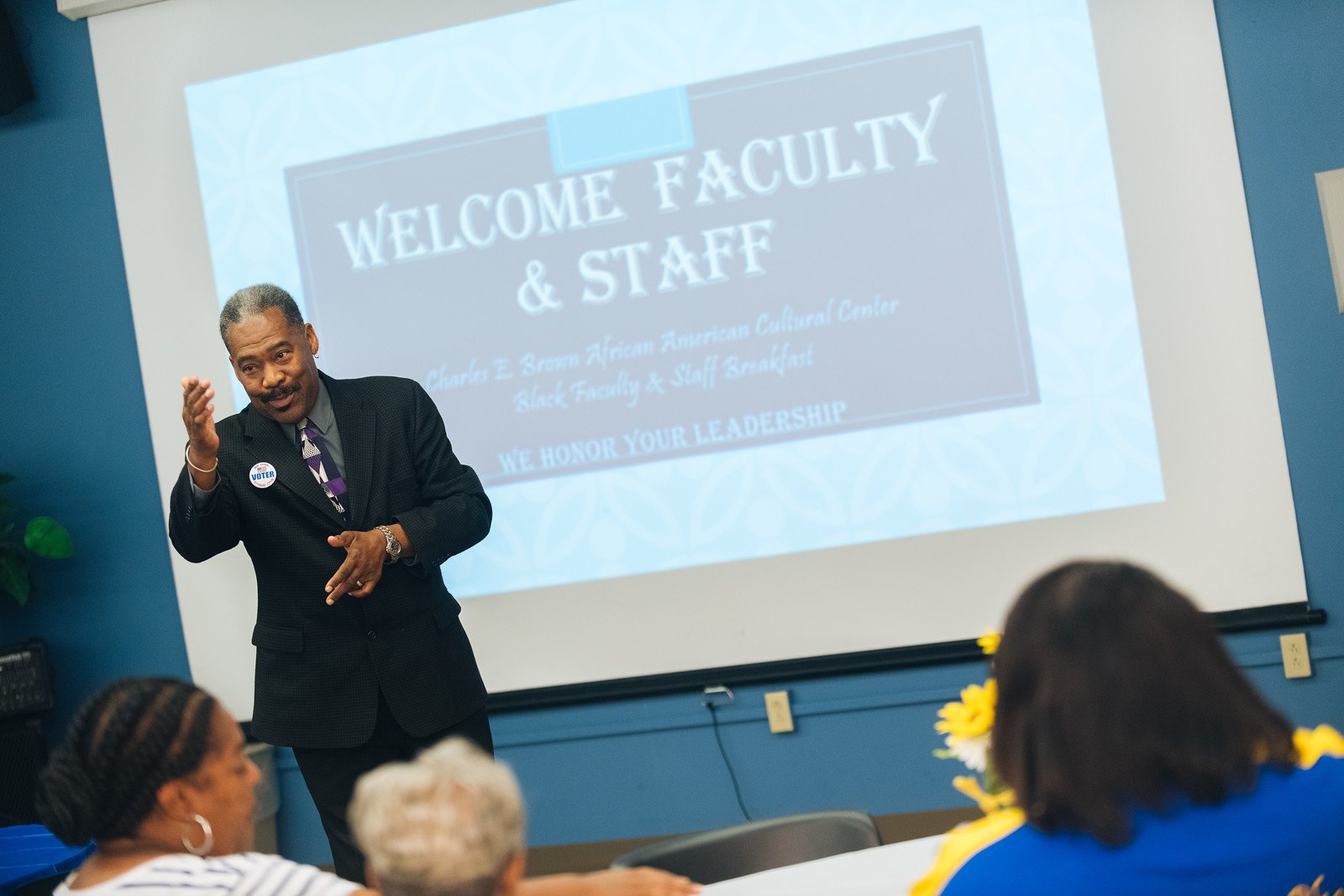 A man in a suit speaks in front of a group of faculty and staff with a presentation slide in the background that says 'Welcome Faculty & Staff