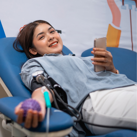 A student smiles while donating blood at the Bleed Blue Blood Drive, holding a stress ball in one hand and a smartphone in the other, lying comfortably on a donation chair.