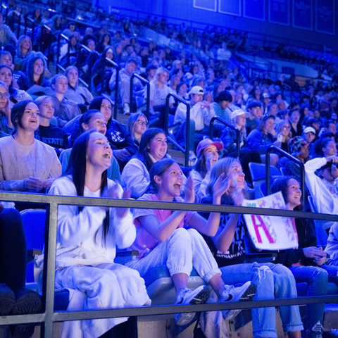 A large crowd of Indiana State University students enthusiastically watches a performance during Entertainment Night, illuminated by blue stage lighting.