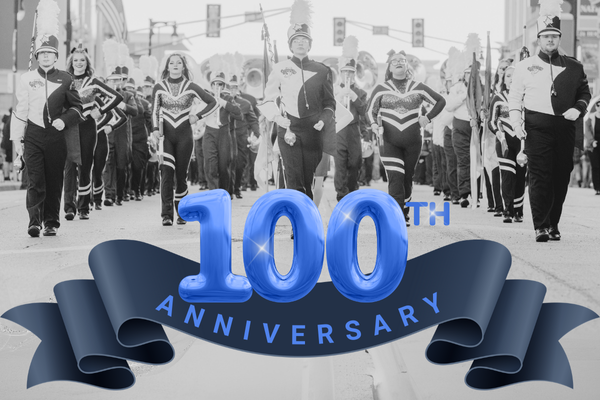 A black-and-white photograph of a marching band performing in a parade, with the text '100th Anniversary' in blue, bold letters overlaid on the image. The number '100' is styled as blue, balloon-like figures, with a blue ribbon beneath them containing the word 'Anniversary.' The band members are wearing traditional marching uniforms with plumes on their hats, and they are marching down a city street.