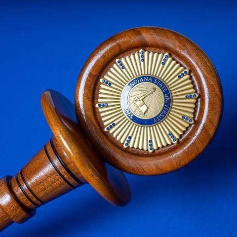 A wooden ceremonial gavel lies against a blue background, with the Indiana State University Alumni Association seal embellished on its striking surface. The seal features a detailed crest, surrounded by a golden circle with numerous small blue gems.