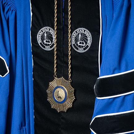 Close-up of a ceremonial academic robe in blue and black, adorned with the Indiana State University seal embroidered on the fabric. A golden medallion featuring the university seal hangs from a chain, symbolizing academic authority and tradition.