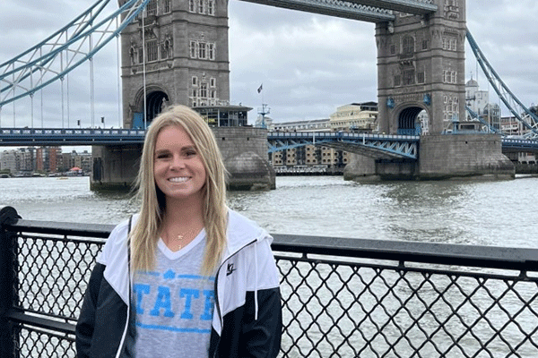 A white female student with straight blond hair poses in front of the London Bridge in London, England. She wears a light grey T-shirt with STATE in blue lettering and a black-and-white rain jacket. A river is visible behind her.