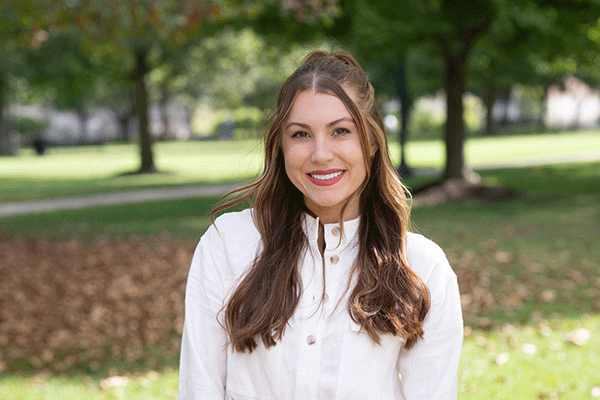 A white female student with wavy brown hair poses outside, wearing a white button-up shirt. Trees and fallen leaves are visible behind her. 