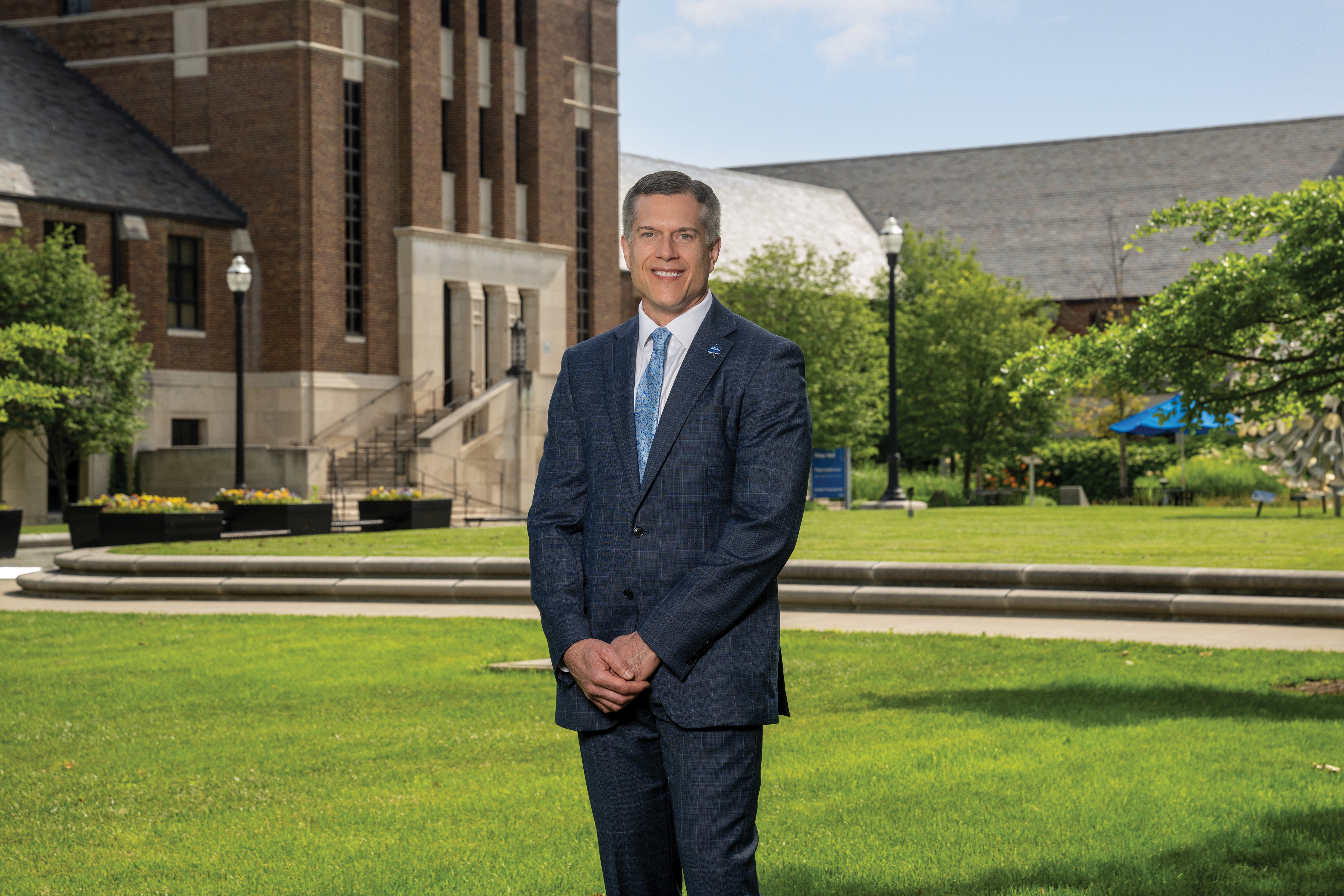 Indiana State University President Mike Godard poses outside with his hands folded. He is a white middle-aged male with greying hair. He wears a dark blue dress suit, a white undershirt, and a light blue tie. Visible behind him is a brick building, a grassy lawn, and other greenery. 