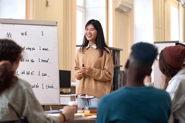 A smiling Asian woman with shoulder-length black hair stands before three peers with a black marker in hand. She wears a white shirt beneath a tan sweater with blue jeans. To her right is a whiteboard with syllables of a language written out and English words – I want, I need, I can, I think – written next to them. Her peers, blurred but visible, are facing her and away from the camera. They are in a brightly lit building space.