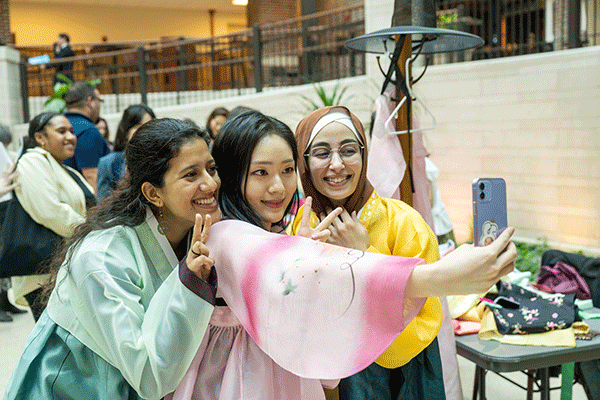 Three female students of diverse international heritage pose for a selfie during a cultural celebration event on campus. They wear colorful, billowing clothing of green, pink, and yellow hues. 