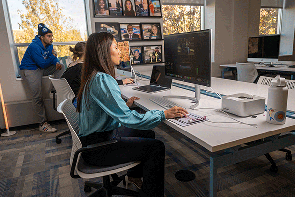 A female student sits in front of a large computer monitor while editing a video. She has dark hair and wears an aqua-blue blouse and dark blue slacks. In the background, another female student is partially visible seated in front of a monitor, while a male student in a blue hoodie and black-and-blue winter cap looks over her shoulder. A laptop sits on the table between the two women. 