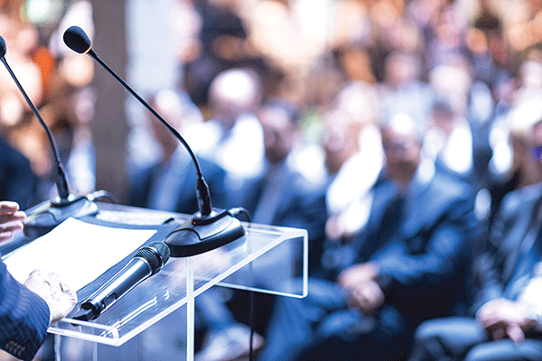 Detailed image of a podium with microphones set up in the left of the frame. A speaker’s hands are visible at the far left. An out-of-focus audience of business professionals is visible in the background.