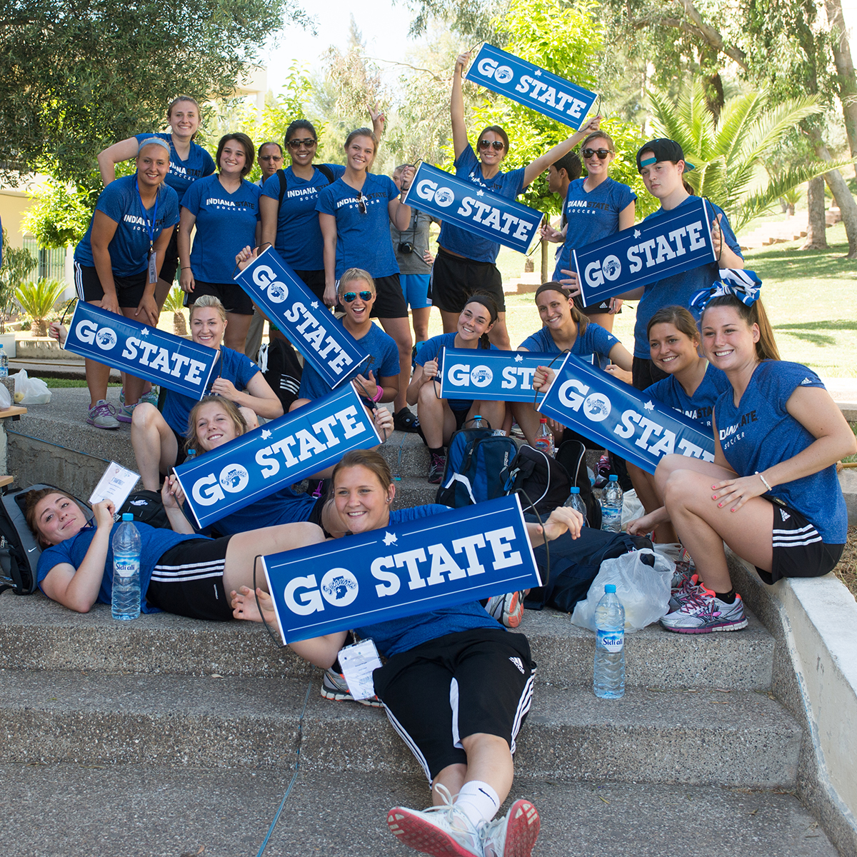 A group of students and staff clad in blue shirts and black pants hold up blue signs with mantras such as "God blue!" printed on them. They are sitting near one another in a stairway on a small hill with tropical vegetation swaying gently in the wind in the background.