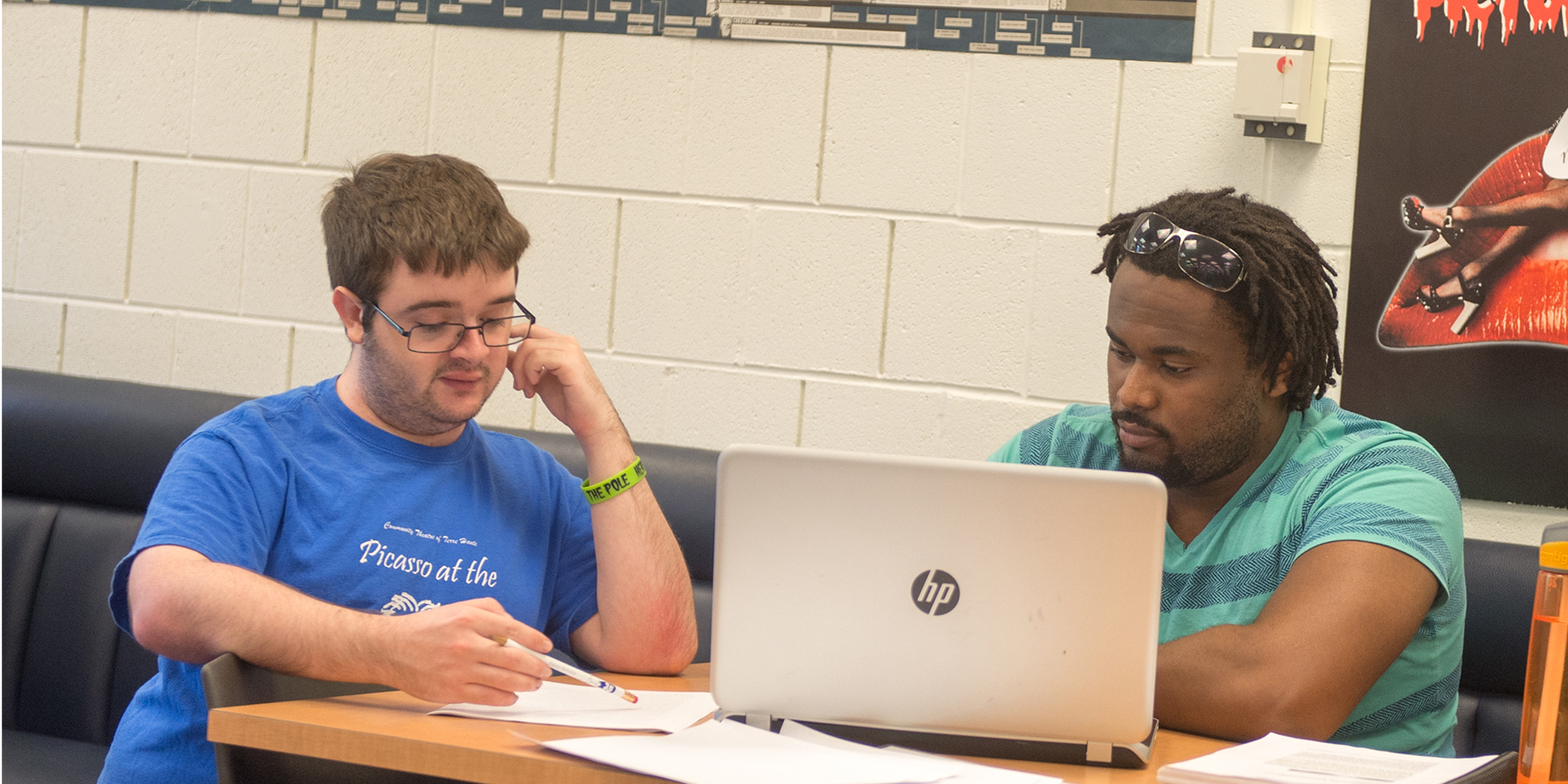 Two students work together in a room with white brick walls and blue benches. The wooden table they are at is covered in papers, with an open HP laptop centered in the frame. They appear to be studying together.