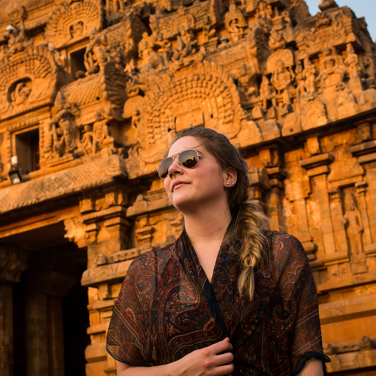 A young woman in aviator sunglasses looks at the setting sun with an ancient temple behind her illuminated orange by the setting sun.