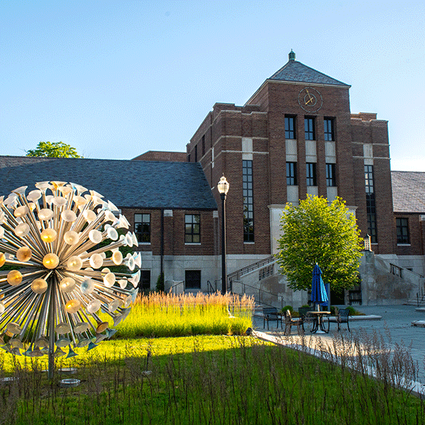 Outdoor scene featuring a modern metal sculpture resembling a dandelion, with a brick building in the background adorned with a clock tower. The setting includes landscaping with green grass, shrubs, and a tree, as well as outdoor seating shaded by blue umbrellas under a clear blue sky