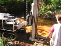 A large piece of large white machinery is set up along the edge of a river. A person stares at it in the corner of the frame wearing a red shirt.