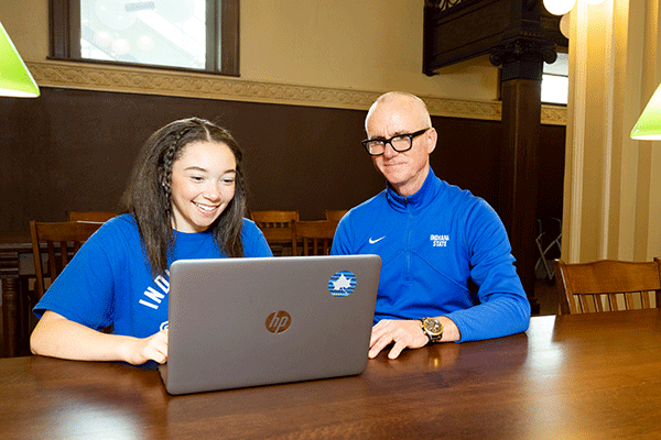 A student and an instructor sit together at a wooden table, smiling while looking at an HP laptop. The student wears a blue 'Indiana State' shirt, and the instructor wears a matching blue Indiana State jacket. The setting appears to be a study area with warm lighting and a classic interior design.