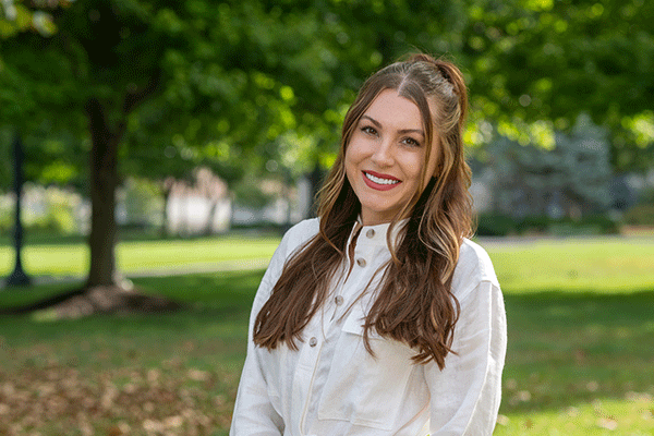 A young female with long brown hair smiling at the camera and standing in the quad in summer with green grass and trees visible.