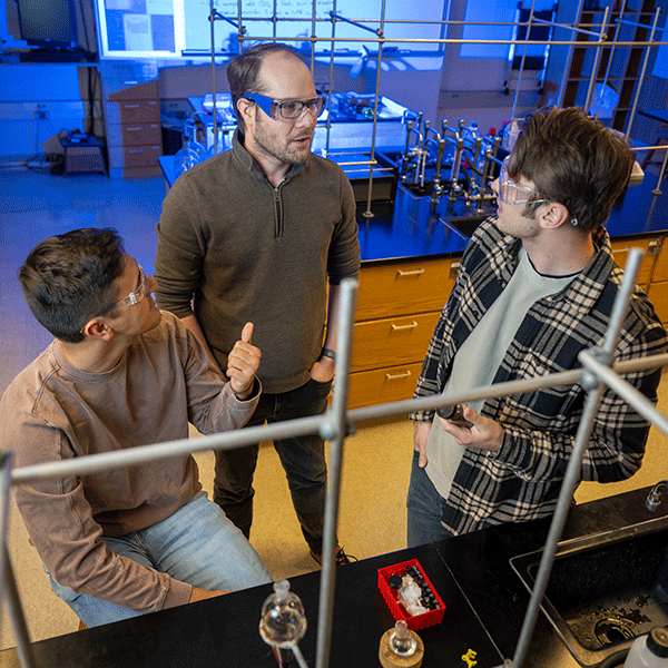 Three individuals in a laboratory setting wearing safety goggles, engaged in a discussion. One person, standing in the center, appears to be instructing or guiding the other two, who are listening attentively. Lab equipment and a sink are visible on the table, with scientific apparatus in the background, indicating a collaborative or educational environment.