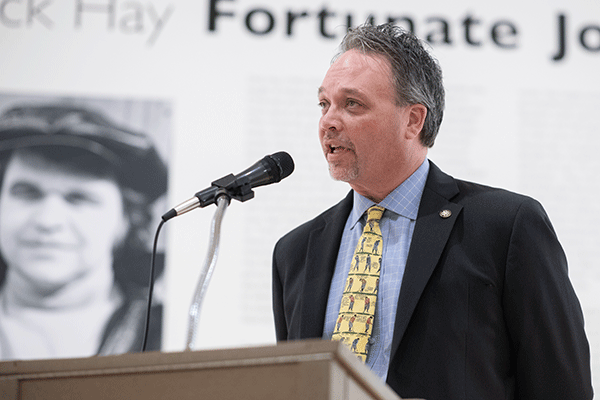 Provost Olsen speaking at a podium with a microphone, dressed in a suit and patterned tie, against a backdrop featuring large text and a black-and-white photograph. The setting appears to be a formal or commemorative event