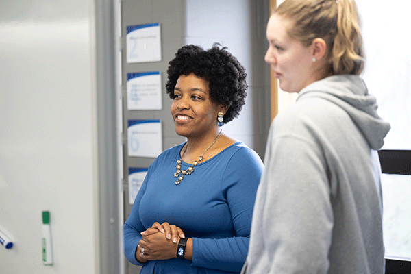 Two women standing in a classroom or office setting, engaged in conversation. The woman on the left, wearing a blue dress and a necklace, smiles warmly, while the woman on the right, dressed casually in a gray hoodie, listens attentively. A whiteboard with notes or signs is visible in the background, suggesting an educational or professional environment