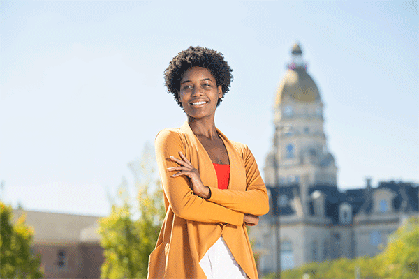 A smiling, tall Black female with short, dark, curly hair stands outside on a sunny day with the Vigo County Courthouse visible but slightly out of focus behind her. She has her arms crossed and wears a long, burnt-orange jacket over a red t-shirt and a white skirt. Trees and greenery are also visible around the courthouse and other buildings behind her.  