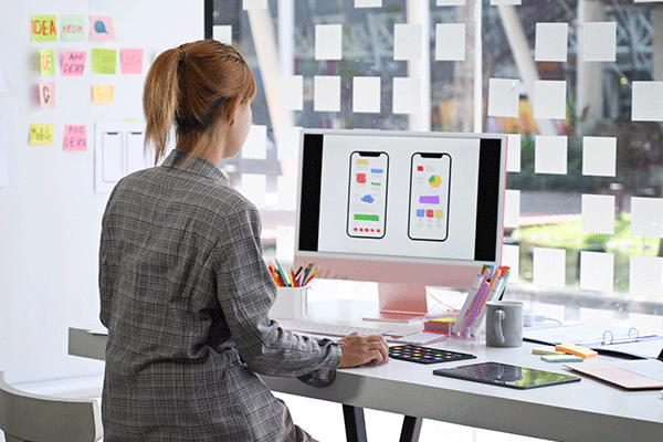 Router_design – A female with her back to the camera sitting in front of a large computer monitor looking at a graphic design with Post-It notes posted on a large window in front of the screen.