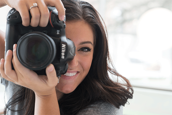 A young female holding a large Nikon camera vertically while looking through the eye piece with the lens the viewer.