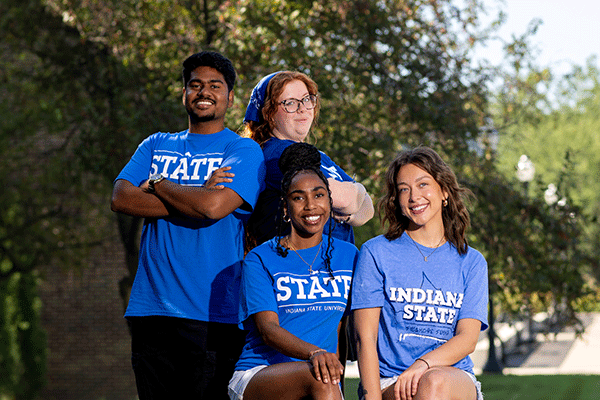 Four students – three female and one male – pose for a group photo, with each one wearing a blue Indiana State University shirt outside in the summer.