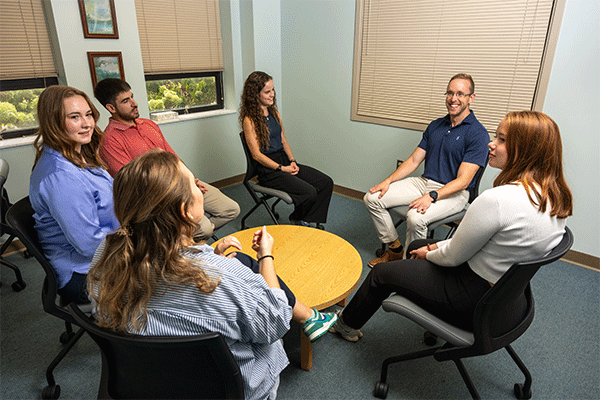 A group of six smiling individuals, a mix of men and women, sits chatting around a small yellow table in a cozy treatment space. They are dressed casually in a variety of styles and colors. There are two windows to the left where greenery can be seen beneath the partially drawn blinds. A larger window to the right has the blinds fully drawn.